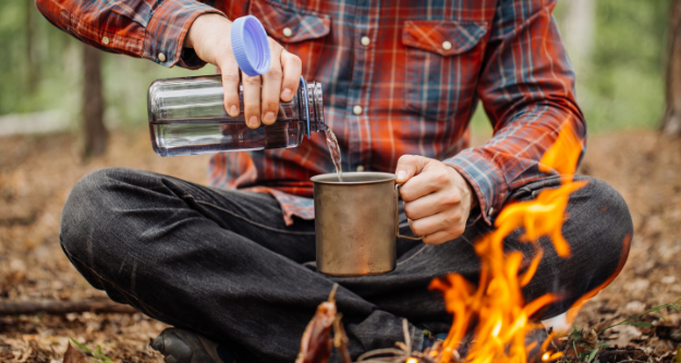 Man in the forest by a fire, poring some water in his cup. - StayWild Outdoor