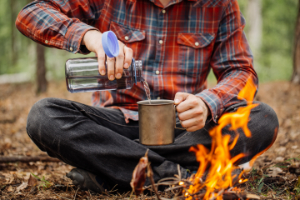 Man in the forest by a fire, poring some water in his cup. - StayWild Outdoor